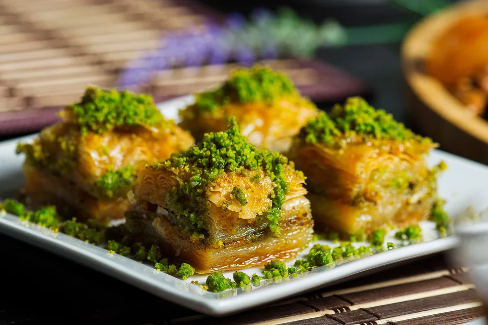 BookNoFees: Foto de ejemplo: Cuatro porciones de baklava en capas, espolvoreadas con pistachos picados, en un plato blanco